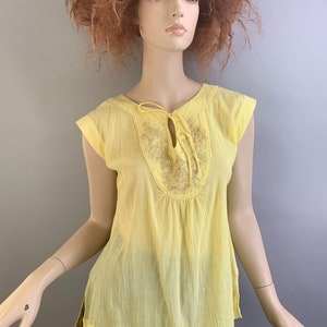 May include: A yellow, semi-sheer, short-sleeved blouse with a tie at the neckline and embroidered detail. The blouse has a gathered waist and a slightly longer back hem.