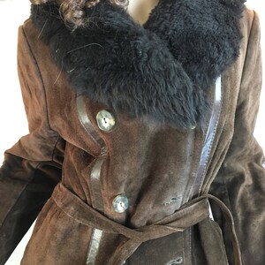 May include: A brown suede coat with a black fur collar and a matching belt. The coat features large, round buttons and leather trim. The coat is a double-breasted style and has a classic, vintage look.