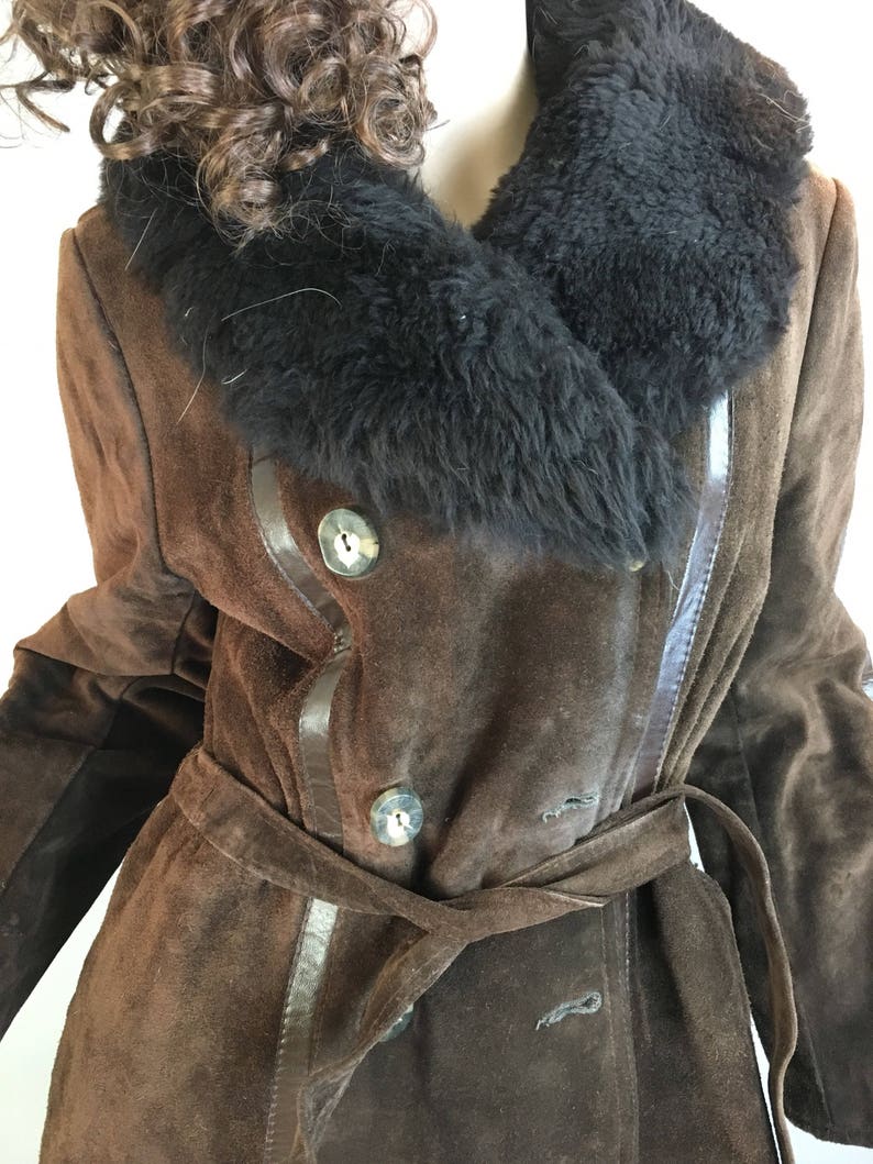 May include: A brown suede coat with a black fur collar and a matching belt. The coat features large, round buttons and leather trim. The coat is a double-breasted style and has a classic, vintage look.