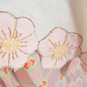 May include: A white linen tablecloth with a pink and brown embroidered floral design. The tablecloth has a scalloped edge and is decorated with pink flowers with brown centers.
