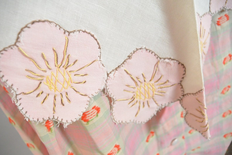 May include: A white linen tablecloth with a pink and brown embroidered floral design. The tablecloth has a scalloped edge and is decorated with pink flowers with brown centers.