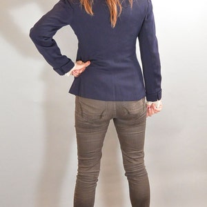 May include: A woman wearing a dark blue blazer, brown jeans, and brown heeled sandals. She is standing with her back to the camera.