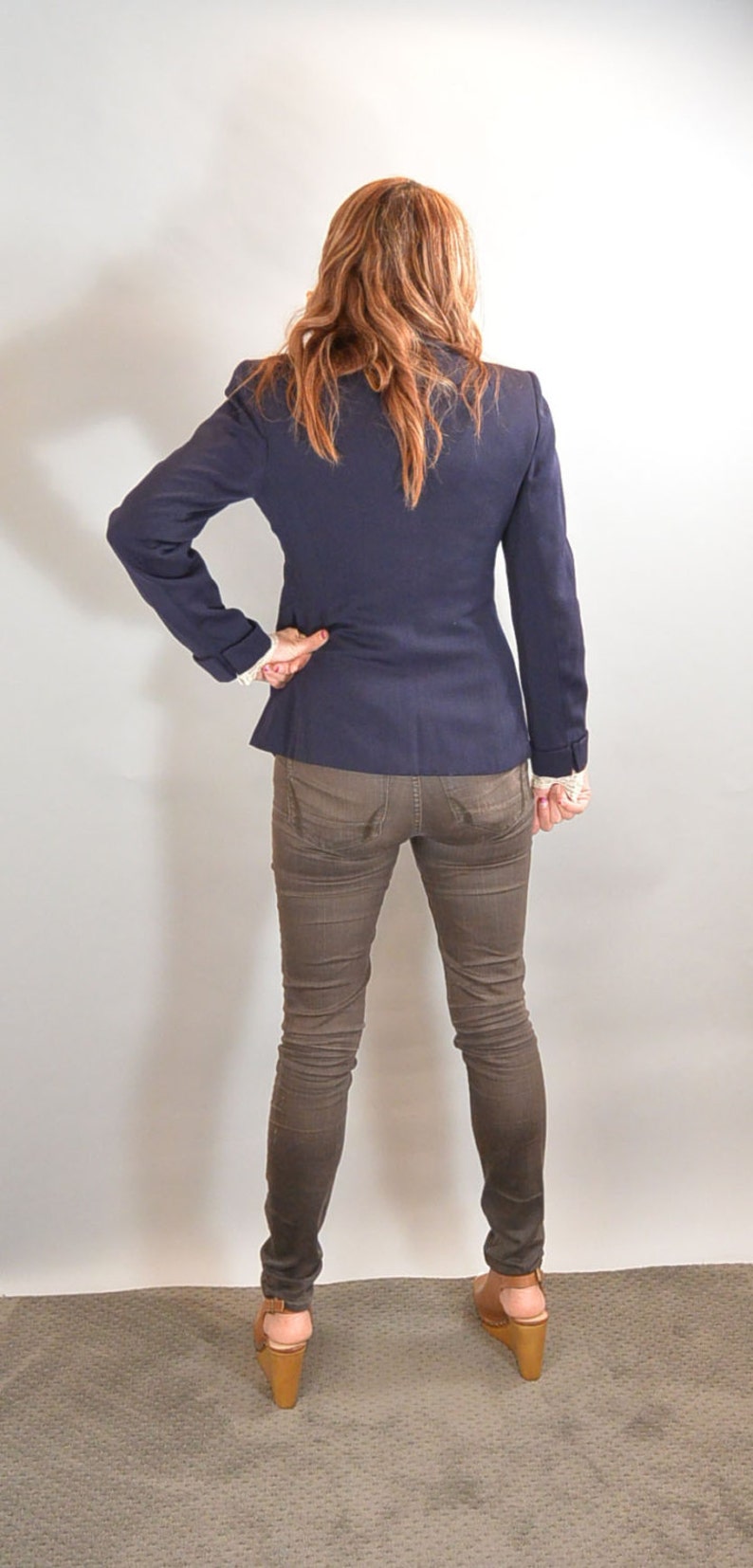 May include: A woman wearing a dark blue blazer, brown jeans, and brown heeled sandals. She is standing with her back to the camera.
