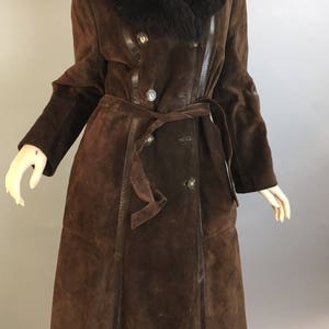 May include: A long, brown suede coat with a black fur collar and a matching belt. The coat features a double-breasted design with large buttons and leather trim. The coat is displayed on a mannequin.