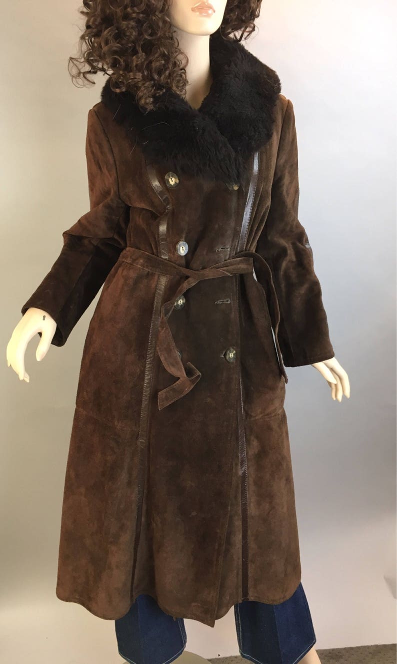 May include: A long, brown suede coat with a black fur collar and a matching belt. The coat features a double-breasted design with large buttons and leather trim. The coat is displayed on a mannequin.