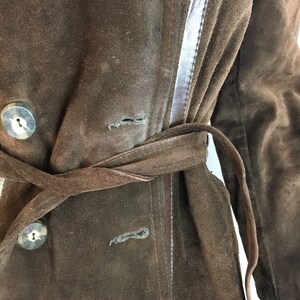 May include: A brown suede coat with a belted waist and large, round buttons. The coat features a double-breasted design and a contrasting leather trim along the seams. The coat's color is a rich, dark brown.