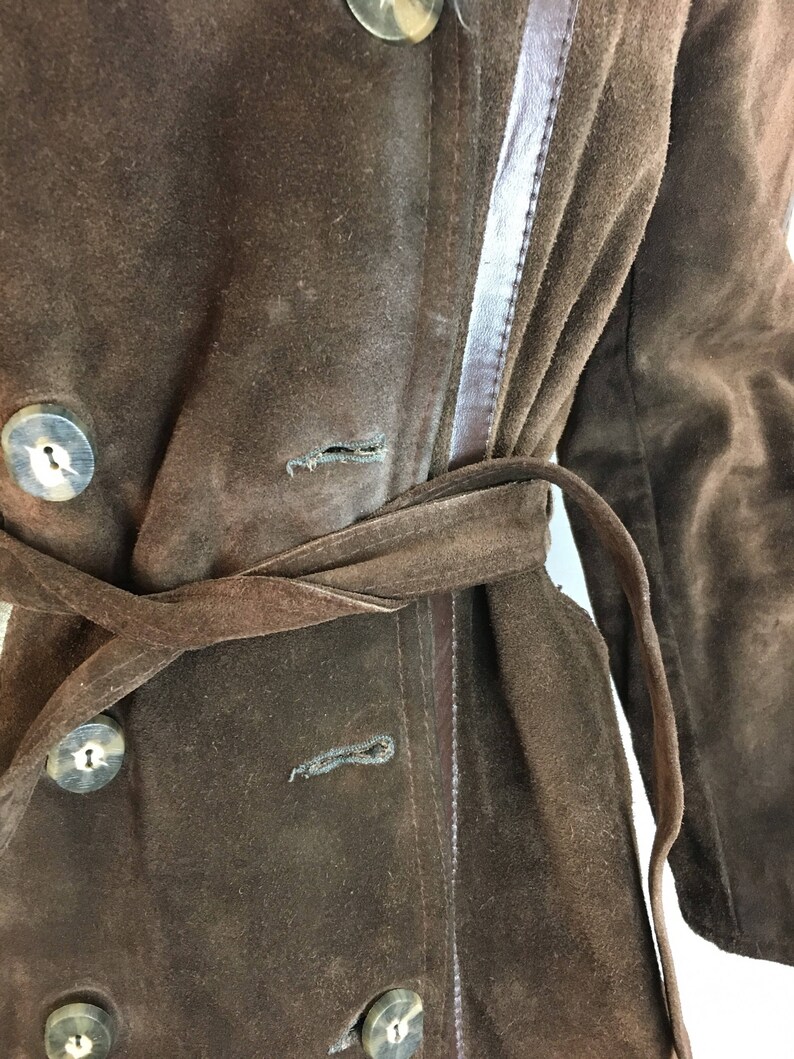May include: A brown suede coat with a belted waist and large, round buttons. The coat features a double-breasted design and a contrasting leather trim along the seams. The coat's color is a rich, dark brown.