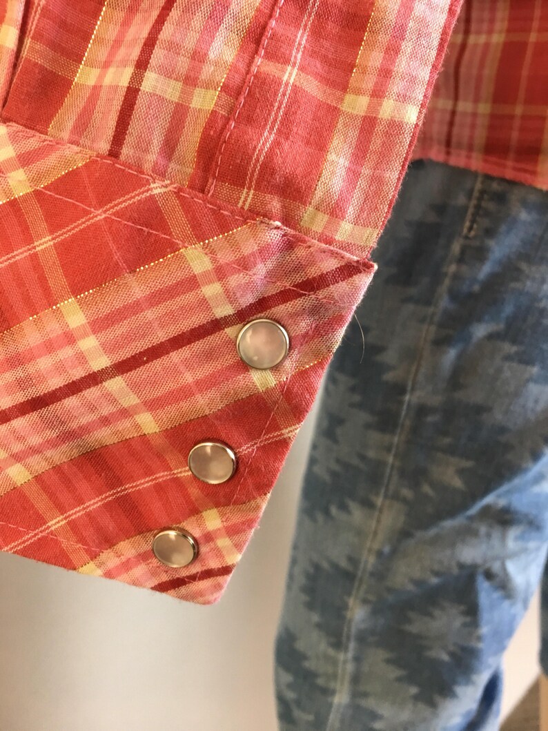 May include: Close-up of a red and yellow plaid shirt cuff with three silver snaps.