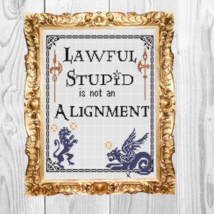 May include: Cross-stitch art in a gold ornate frame. The text reads "Lawful Stupid is not an Alignment." The design includes a lion and a griffin. The background is a white wooden surface.