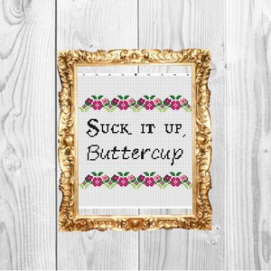 May include: A framed cross-stitch design with the text "Suck it up, Buttercup" in black lettering. The design features a border of pink and green flowers.