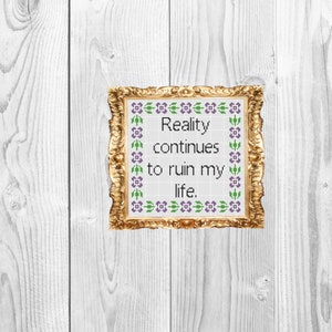 May include: A gold framed picture with a white background and purple and green floral border. The text inside the frame reads "Reality continues to ruin my life."