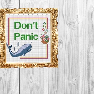 May include: A framed cross-stitch design with a blue whale jumping out of a pot of flowers. The text "Don't Panic" is stitched in green letters.