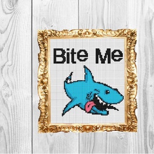 May include: A framed cross-stitch design featuring a blue cartoon shark with a pink tongue sticking out and the text "Bite Me" in black letters.