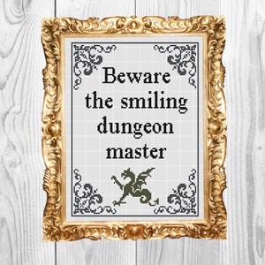 May include: A framed cross-stitch design with a gold frame. The design features a black and white illustration of a dragon with the text "Beware the smiling dungeon master".