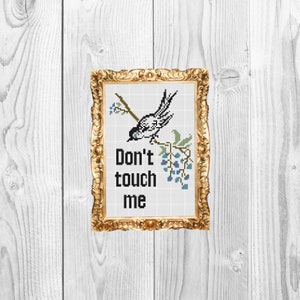May include: A framed cross-stitch design featuring a black and white bird perched on a branch with blue berries. The text "Don't touch me" is below the bird.