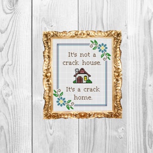 May include: Cross-stitch artwork in a gold frame. The text reads, "It's not a crack house. It's a crack home." The design includes a house illustration and floral accents in blue and green, perfect for home decor.