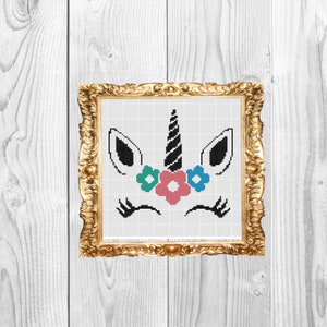 May include: A framed cross-stitch design of a unicorn face with a black horn, black eyes, and a pink, blue, and green flower crown.
