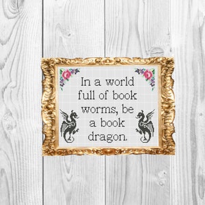 May include: A framed cross-stitch with a white background and black text that reads "In a world full of book worms, be a book dragon." The frame is gold with a floral design.