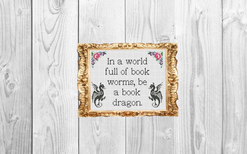 May include: A framed cross-stitch with a white background and black text that reads "In a world full of book worms, be a book dragon." The frame is gold with a floral design.