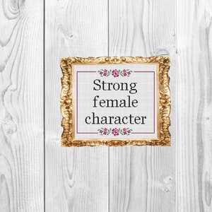 Op de afbeelding: Een gouden ingelijst beeld met een witte achtergrond en de tekst "Strong female character" in zwarte letters. De tekst wordt omringd door een bloemmotief met roze bloemen.