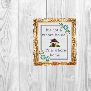 May include: Cross-stitch art in a gold ornate frame. The artwork features the text "It's not a whore house. It's a whore home." A small house graphic is in the center. Blue flowers and green leaves decorate the corners.