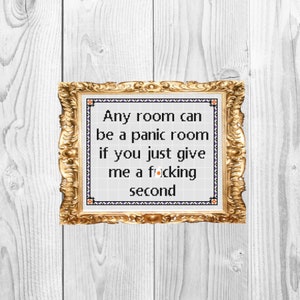 Op de afbeelding: Een ingelijste kruissteek met de tekst "Any room can be a panic room if you just give me a f*cking second." De gouden lijst heeft sierlijke details. De achtergrond is een wit houten oppervlak.