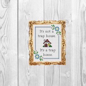 Puede incluir: Obra de arte de punto de cruz en un marco dorado ornamentado. La obra presenta una casa con tejado rojo y el texto "It's not a trap house. It's a trap home." Acentos florales azules adornan las esquinas. El fondo es de vetas de madera blanca.