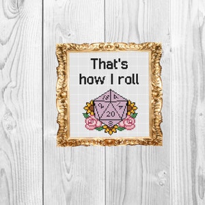 Może przedstawiać: Ramka z haftem krzyżykowym przedstawiająca fioletową 20-ścienną kostkę do gry z tekstem "That's how I roll". Kostka otoczona jest różowymi kwiatami i żółtymi słonecznikami.