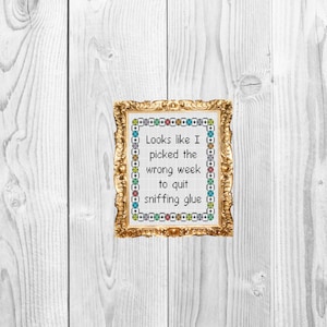 May include: A framed cross-stitch design with a gold frame. The cross-stitch design features a colorful border and the text "Looks like I picked the wrong week to quit sniffing glue".