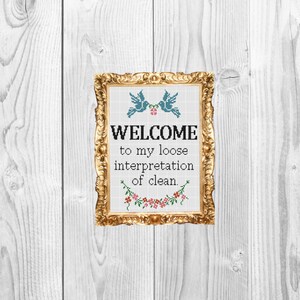 Könnte beinhalten: Ein goldfarbener Kreuzstichrahmen mit blauen Vögeln und rosa Blumen. Der Text lautet "WELCOME to my loose interpretation of clean."