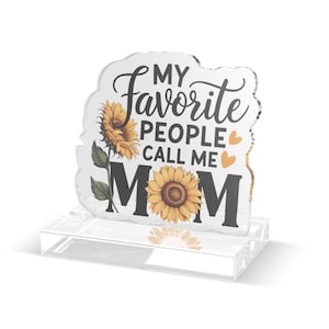 Może przedstawiać: Przezroczysty blok akrylowy z napisem "MY Favorite PEOPLE CALL ME MOM" w czarnym skrypcie i blokowych literach. Projekt zawiera żółte słoneczniki i pomarańczowe serca. Blok stoi na przezroczystej podstawie.