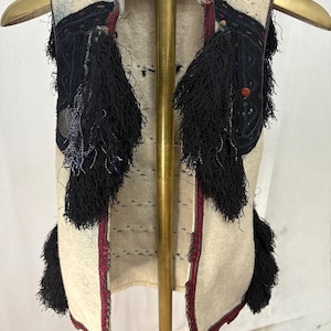 以下が含まれることがあります： 黒い刺繍と長い黒いタッセルが付いた、クリーム色のベスト。 ベストの縁には赤いトリムがあり、金色のハンガーに掛けられています。 ベストは厚手のテクスチャード加工の生地で作られているようです。
