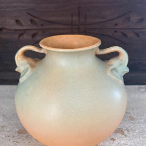 May include: A ceramic vase with a rounded body and two handles. The vase has a gradient color scheme, transitioning from a light green at the top to a peach color at the base. The interior and handles are a light brown color.