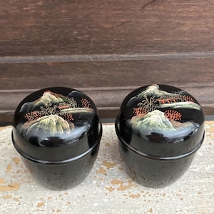 May include: Two black, round, lidded boxes with a landscape design on the lids. The landscape features a mountain range, trees, and a building, all in shades of white, orange, and green. The boxes are sitting on a light-colored surface.