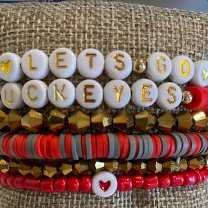 May include: A stack of five beaded bracelets featuring white round beads with gold lettering that spells LETS GO BUCKEYES, interspersed with red, grey, and metallic gold faceted beads. The design includes small heart accents and gold spacer beads.