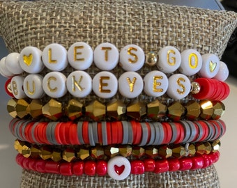 May include: A stack of five beaded bracelets featuring white round beads with gold lettering that spells LETS GO BUCKEYES, interspersed with red, grey, and metallic gold faceted beads. The design includes small heart accents and gold spacer beads.