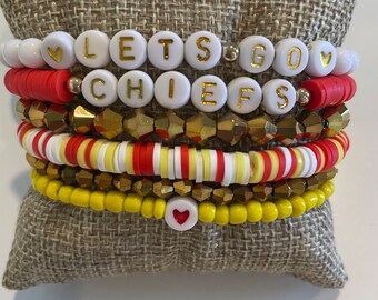 May include: A stack of bracelets with red, white, and yellow beads. The bracelets spell out "Let's Go Chiefs" with gold letters on white beads. There are also small red heart beads.