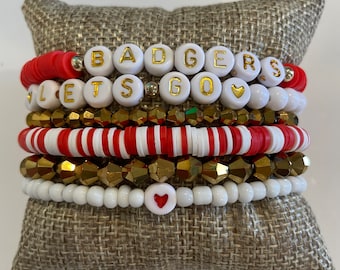 May include: A stack of bracelets with red and white striped beads, gold beads, and white beads with gold lettering spelling "BADGERS LET'S GO" with a heart charm.