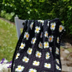 以下が含まれることがあります： 白いデイジーの花が咲いた黒いクロシェの毛布。毛布は白い椅子にかけられており、波打つ縁取りがあります。