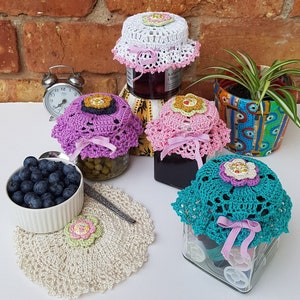 May include: Four glass jars with crocheted lids. The lids are in various colors, including pink, white, and teal. The jars are filled with different items, including olives, pickles, and sewing supplies. The jars are sitting on a table with a white crocheted doily.
