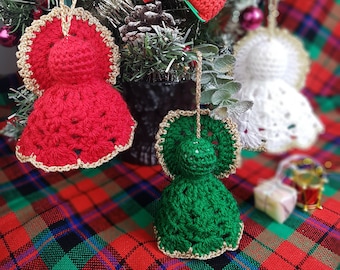 May include: Three crocheted Christmas ornaments: a red angel, a green angel, and a white angel. The ornaments are hanging on a Christmas tree with red and green plaid fabric underneath.