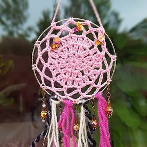 May include: Pink crocheted dream catcher with white, pink, and black yarn tassels and gold beads hanging from a metal ring.