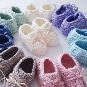 May include: A collection of ten pairs of crocheted baby booties in various colors, including pink, blue, green, white, and gray. Each pair has a satin ribbon tied around the ankle.
