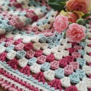 Op de afbeelding: Een gehaakt deken met een granny square patroon in roze, wit en blauw. Het deken heeft een rand met afwisselende strepen roze, wit en blauw. Het deken ligt op een houten oppervlak met roze rozen erop.