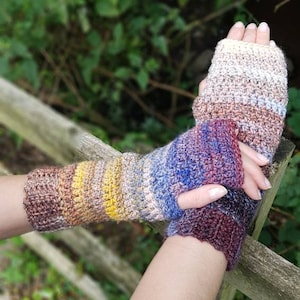 Puede incluir: Un par de guantes sin dedos de ganchillo de colores con un degradado de marrón, amarillo, azul y morado. Los guantes están hechos con un punto texturizado.