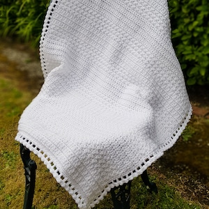May include: A white crocheted baby blanket with a delicate crocheted border. The blanket is draped over a black metal chair in a grassy outdoor setting.