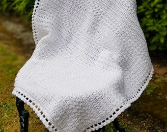 May include: A white crocheted baby blanket with a delicate crocheted border. The blanket is draped over a black metal chair in a grassy outdoor setting.
