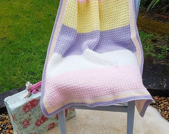 May include: A multi-coloured crocheted blanket with stripes of pink, yellow, white, and purple. The blanket is draped over a white wooden chair.