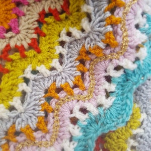 May include: Close-up of a colourful crocheted blanket with a gold chain trim. The blanket is made with different coloured yarn, including pink, blue, yellow, and orange.