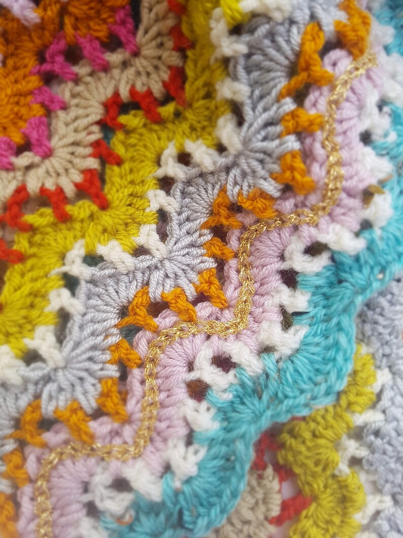 May include: Close-up of a colourful crocheted blanket with a gold chain trim. The blanket is made with different coloured yarn, including pink, blue, yellow, and orange.